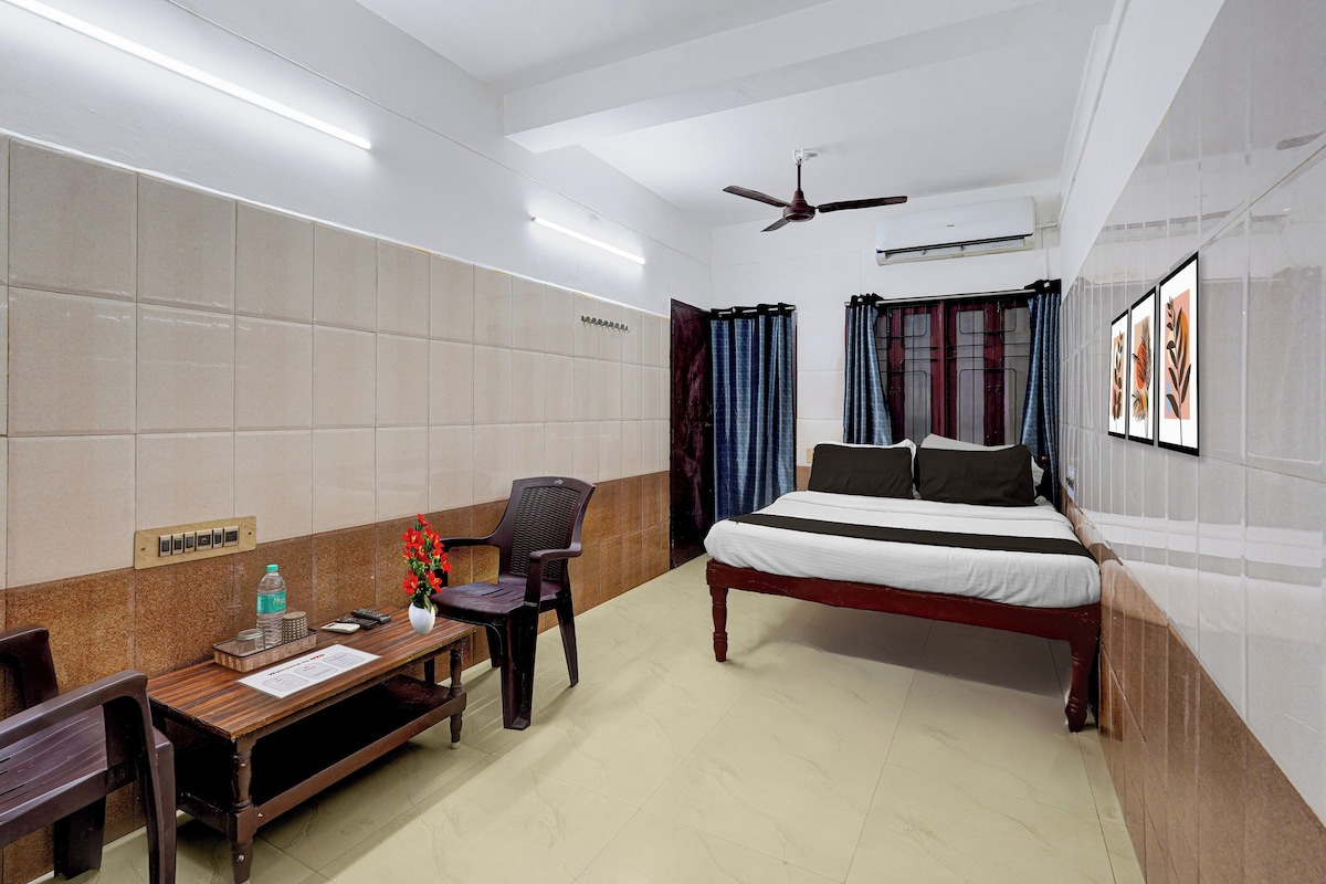 Habitación de hotel con cama, escritorio, silla, botella de agua, jarrón con flores, ventilador de techo y ventana con cortinas.