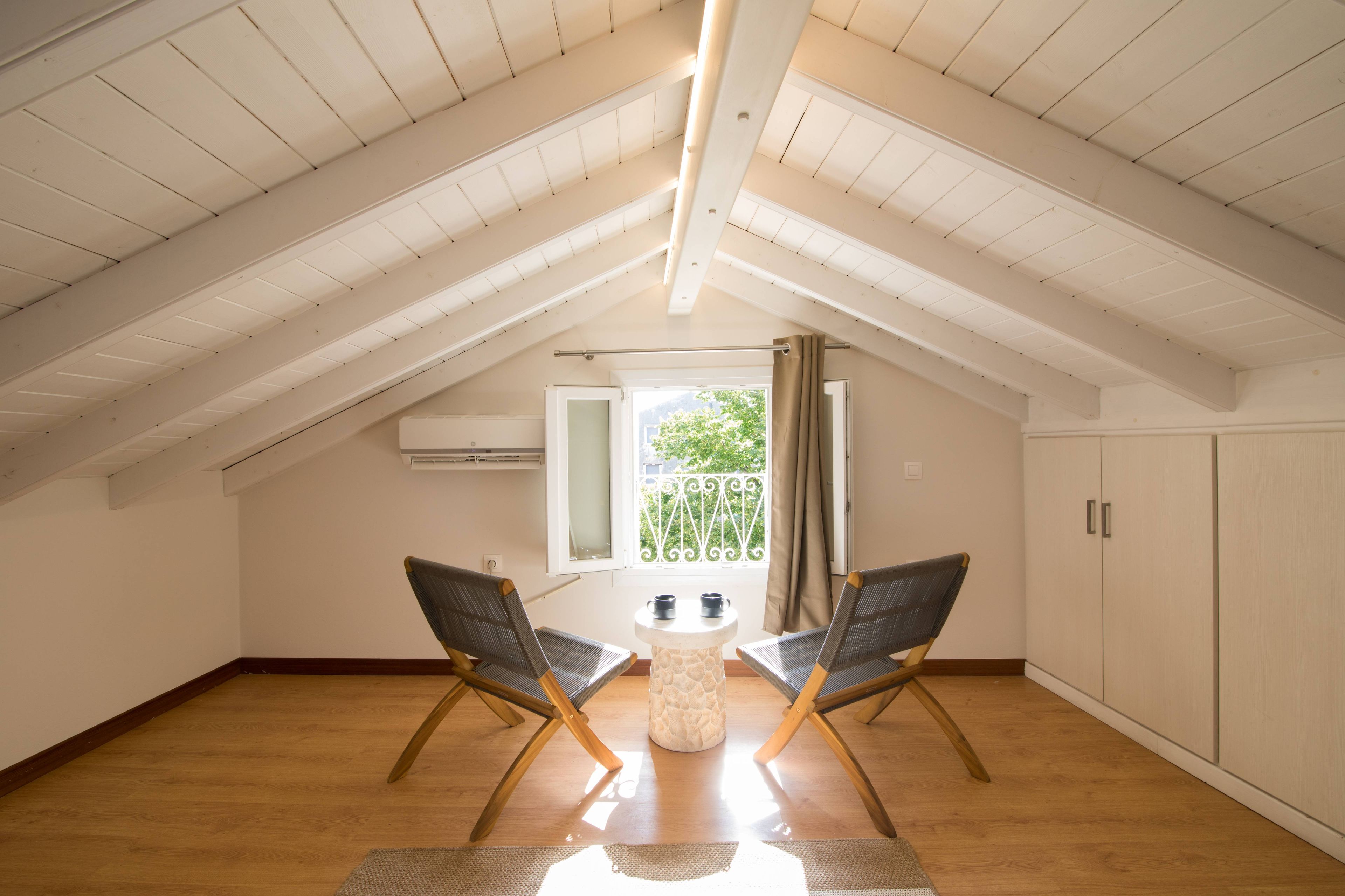 Amplia habitación en el ático con techo inclinado, dos sillas, una mesita y una ventana con vistas a la vegetación.
