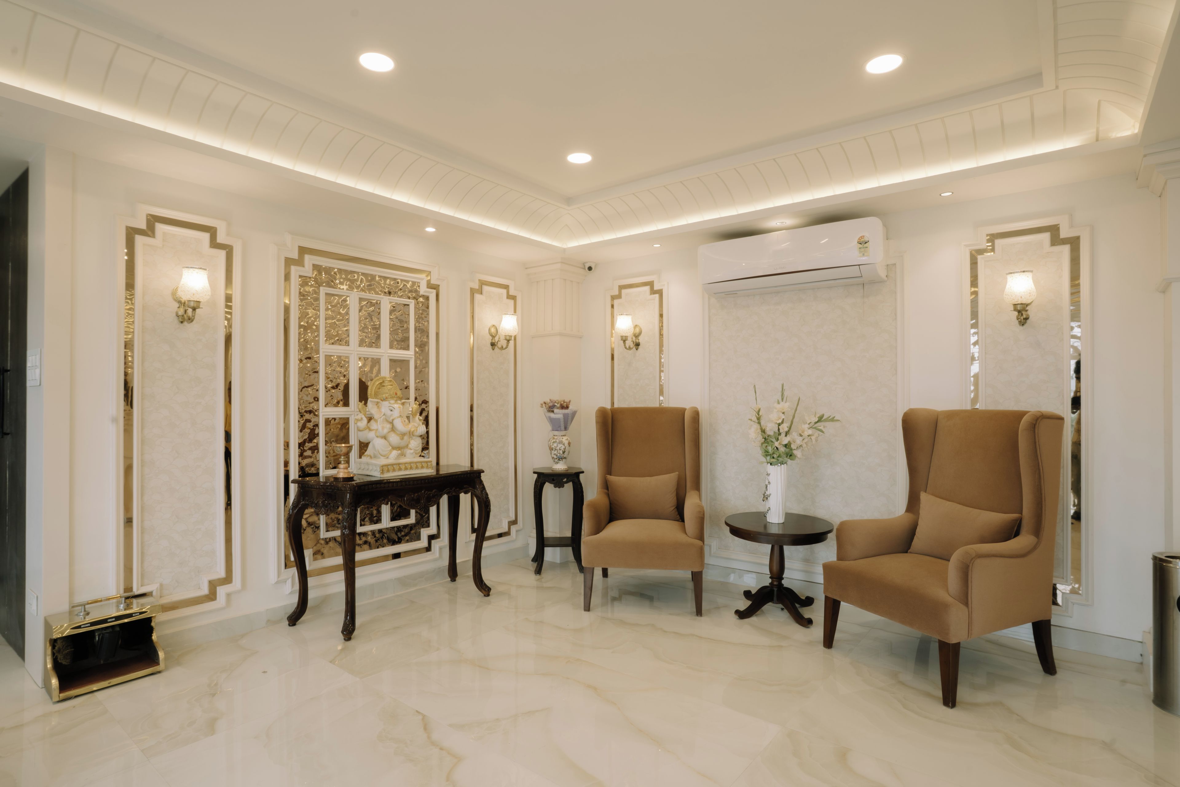 Una habitación amplia con piso de mármol, dos sillones marrones, una mesita redonda y un espejo decorativo con detalles dorados.