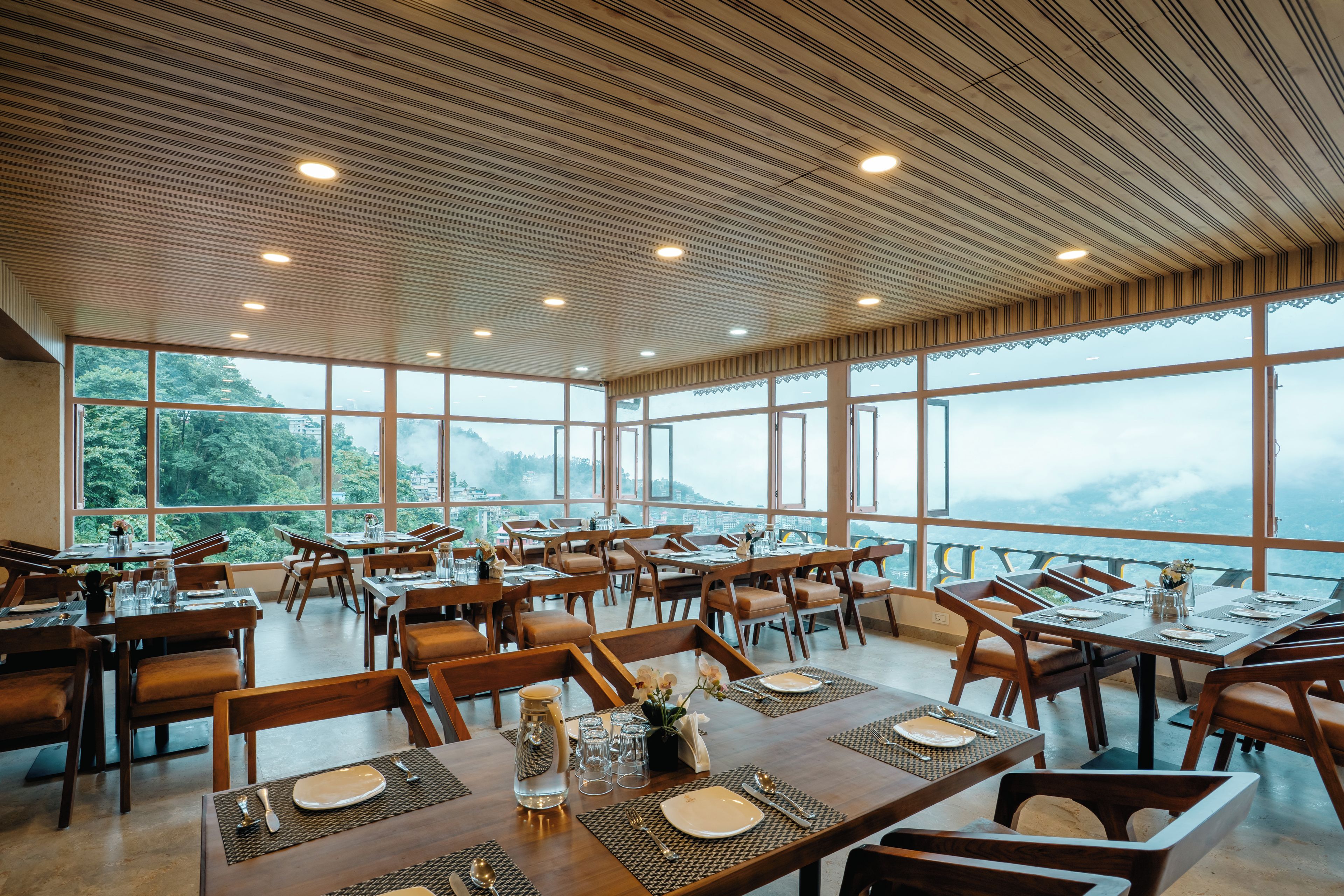 Un restaurante con varias mesas y sillas, amplios ventanales que ofrecen vistas a las montañas y un techo de madera con iluminación empotrada.