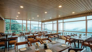 Un restaurante con varias mesas y sillas, amplios ventanales que ofrecen vistas a las montañas y un techo de madera con iluminación empotrada.