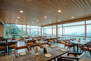 A restaurant with multiple tables and chairs, large windows offering a view of the mountains, and a wooden ceiling with recessed lighting. - Hotel Maneybung (Gangtok)