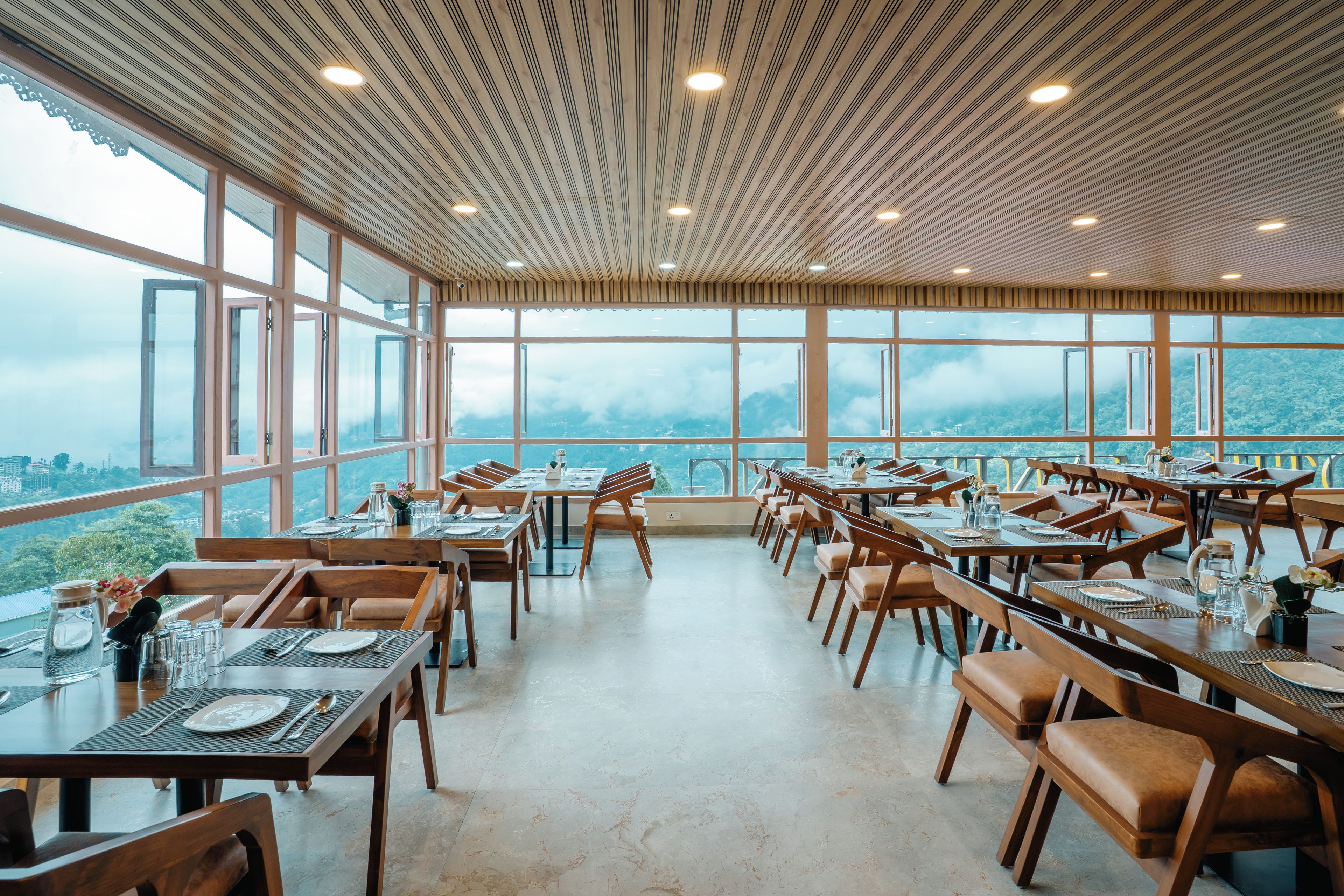 A restaurant with a high ceiling and large windows offering a view of the mountains. Tables are set with plates, cutlery, and glasses.