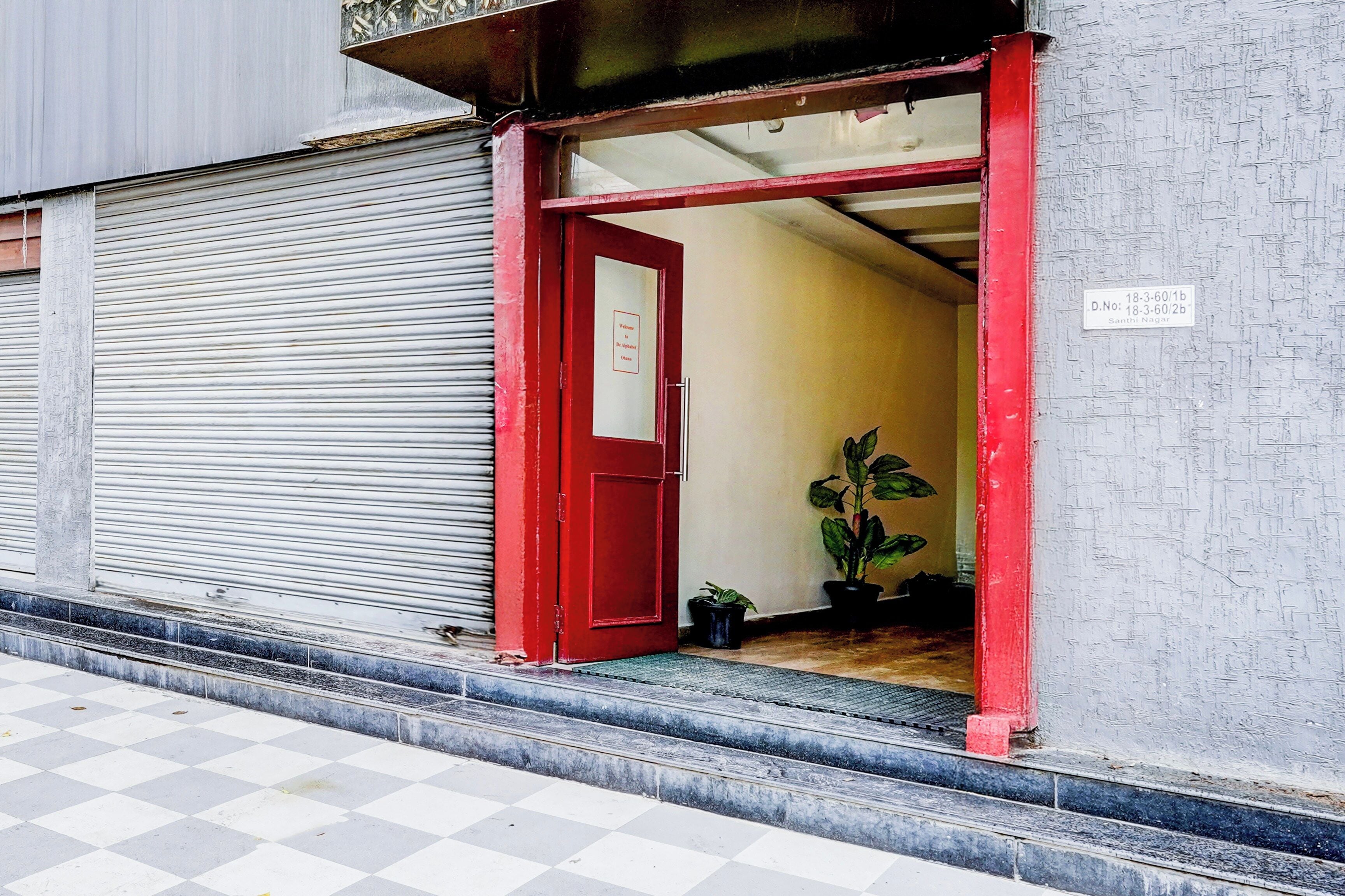 A building entrance with a red door, a closed shutter door, and a potted plant inside.