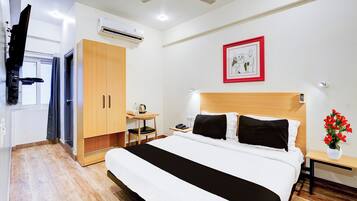 Habitación de hotel con cama, escritorio, aire acondicionado, ventilador de techo, un cuadro enmarco y un florero con flores.