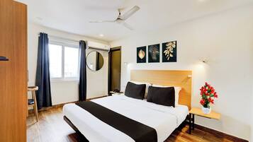 Habitación de hotel con cama, mesita de noche con lámpara, ventilador de techo, ventana con cortinas y una obra de arte en la pared.