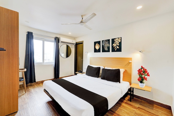 A hotel room with a bed, a bedside table with a lamp, a ceiling fan, a window with curtains, and artwork on the wall.