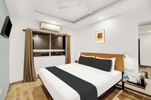 Habitación de hotel con cama, mesita de noche con hervidor, cuadro enmarcado en la pared y ventana con cortinas.