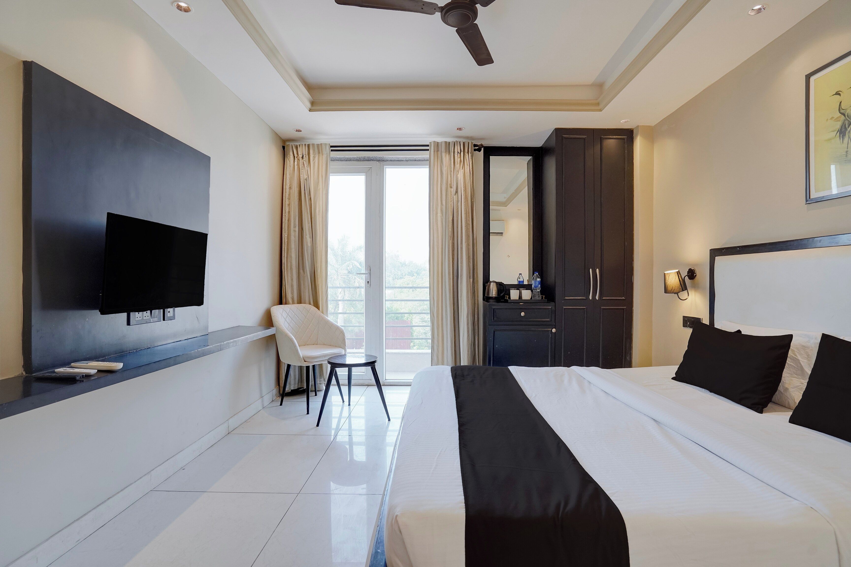 Habitación de hotel con cama, televisor montado en la pared, escritorio con silla, armario y balcón con cortinas.