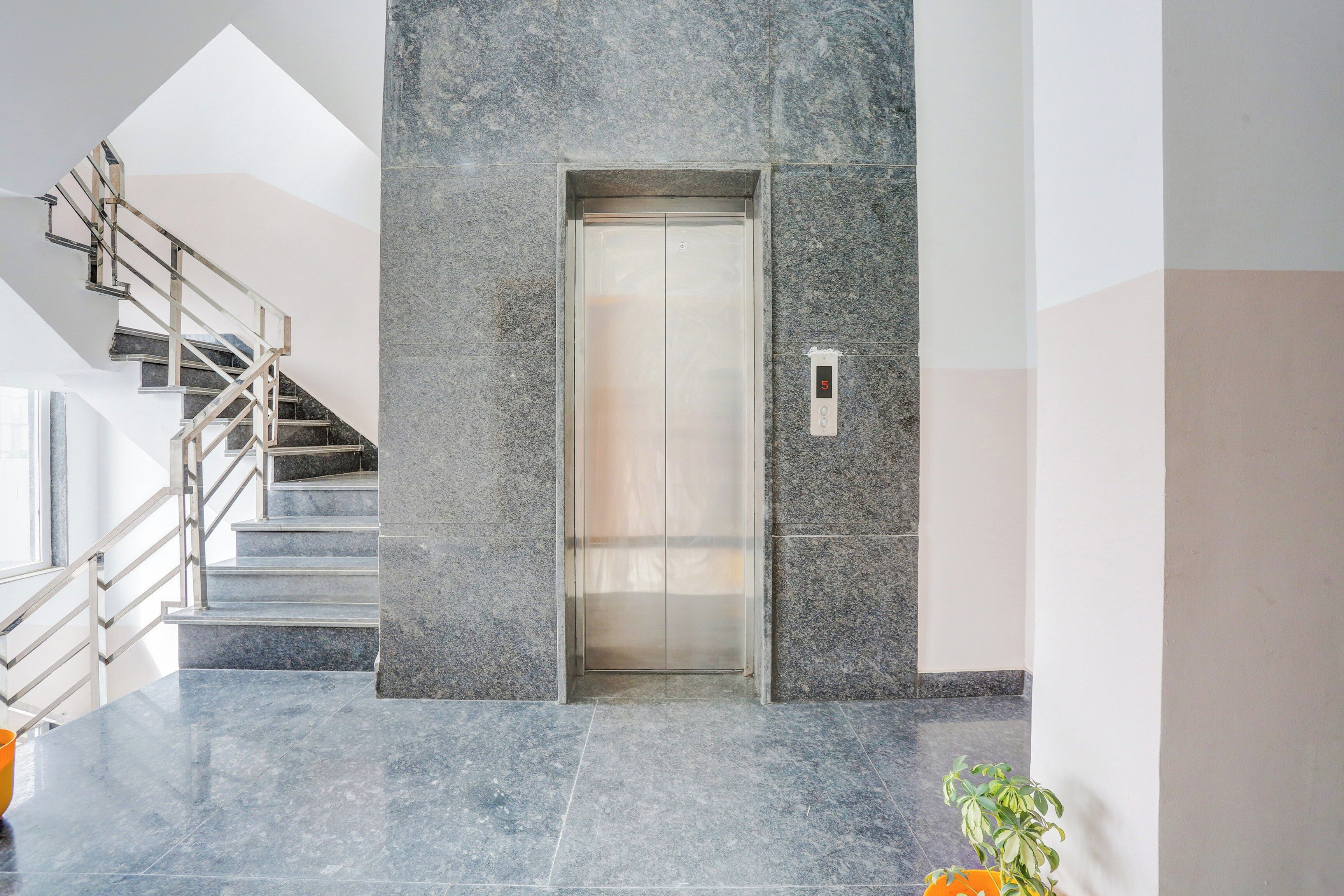 A modern elevator with a digital display, a marble-tiled wall, and a staircase with metal railings.