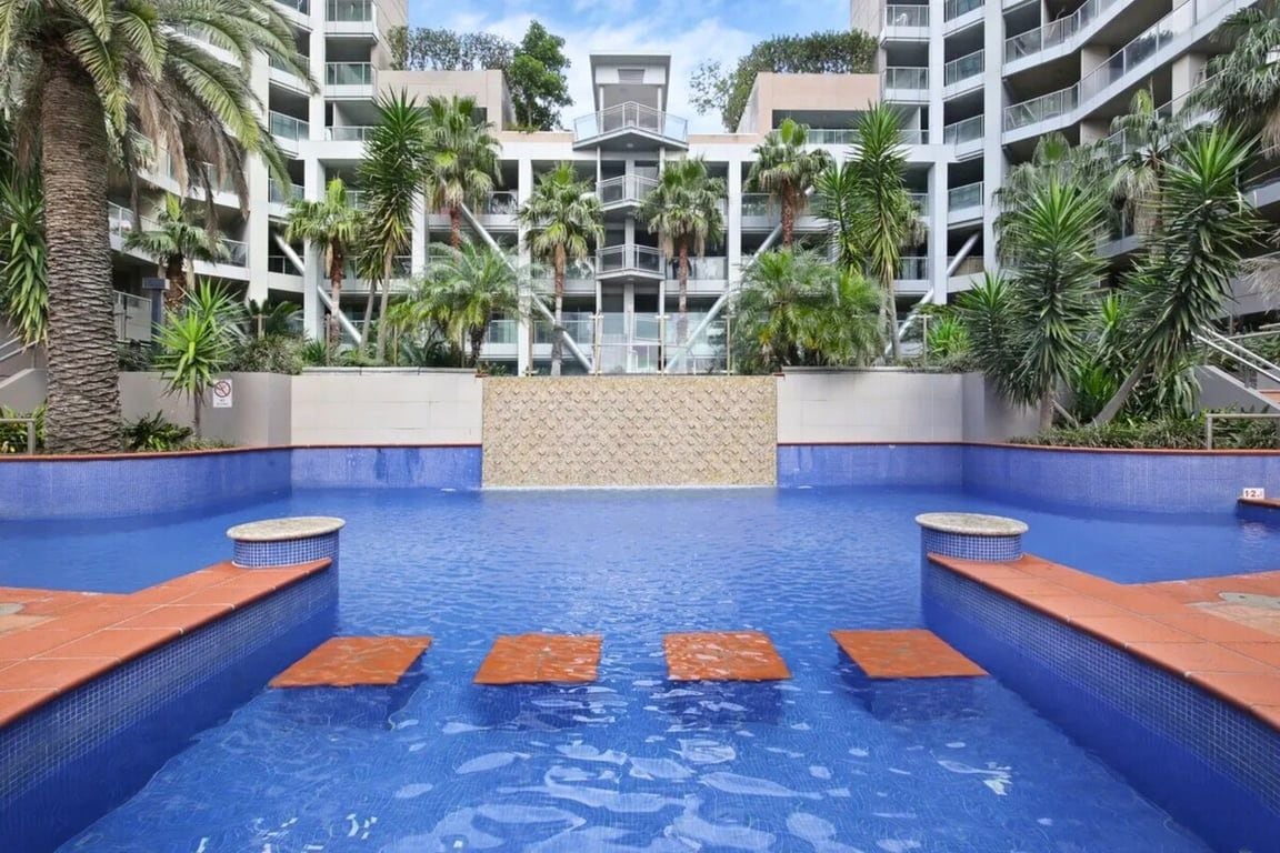 A swimming pool with a blue tiled floor, surrounded by palm trees and a multi-story building with balconies.