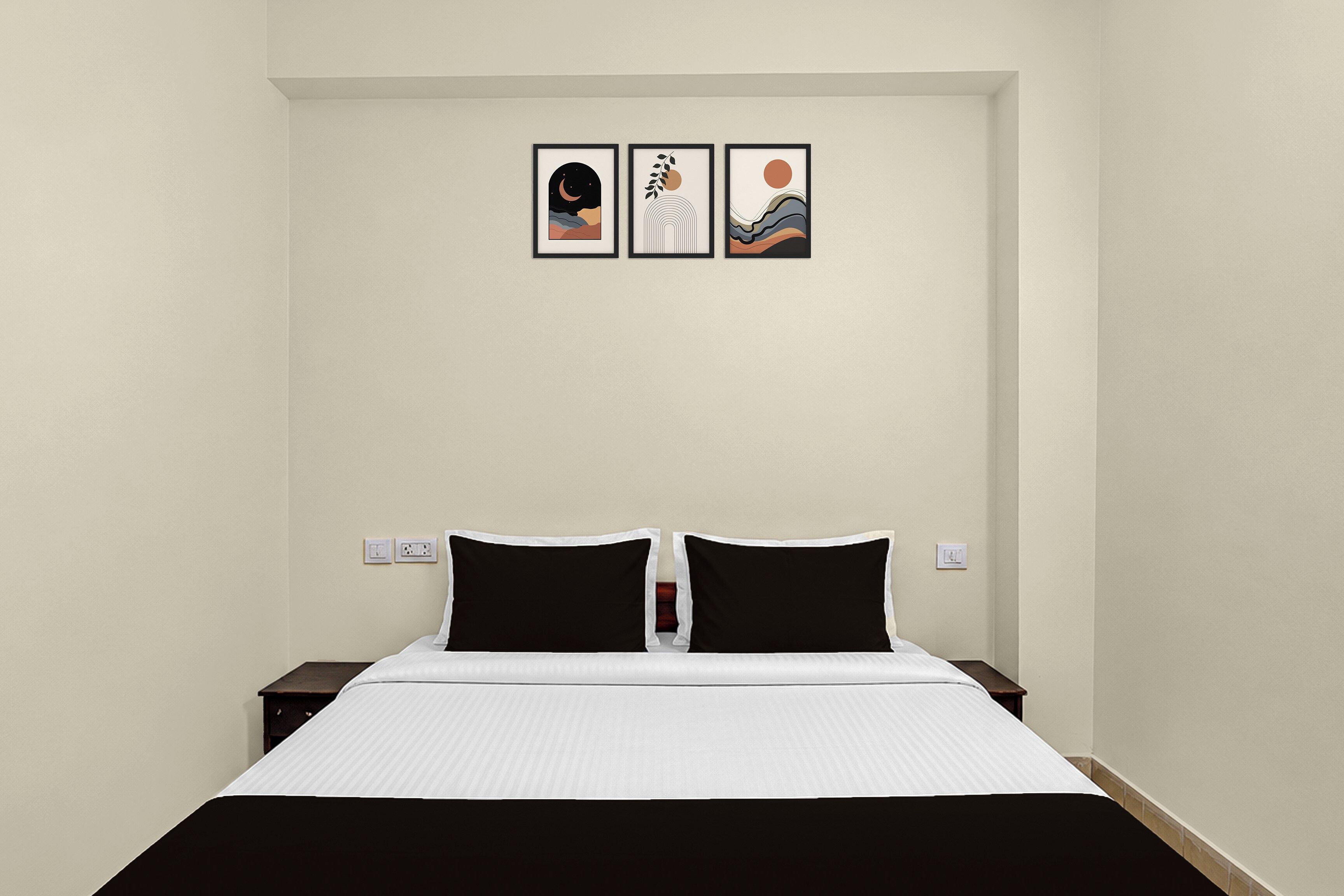 Habitación de hotel con cama, dos almohadas negras, una colcha blanca y tres cuadros enmarcados en la pared.