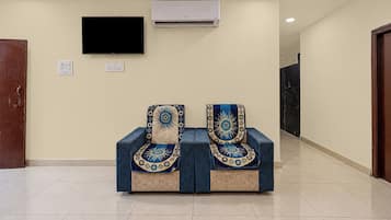 Un pasillo con dos sillones tapizados en azul y cubiertas estampadas, un televisor de pantalla plana montado en la pared y una unidad de aire acondicionado encima.