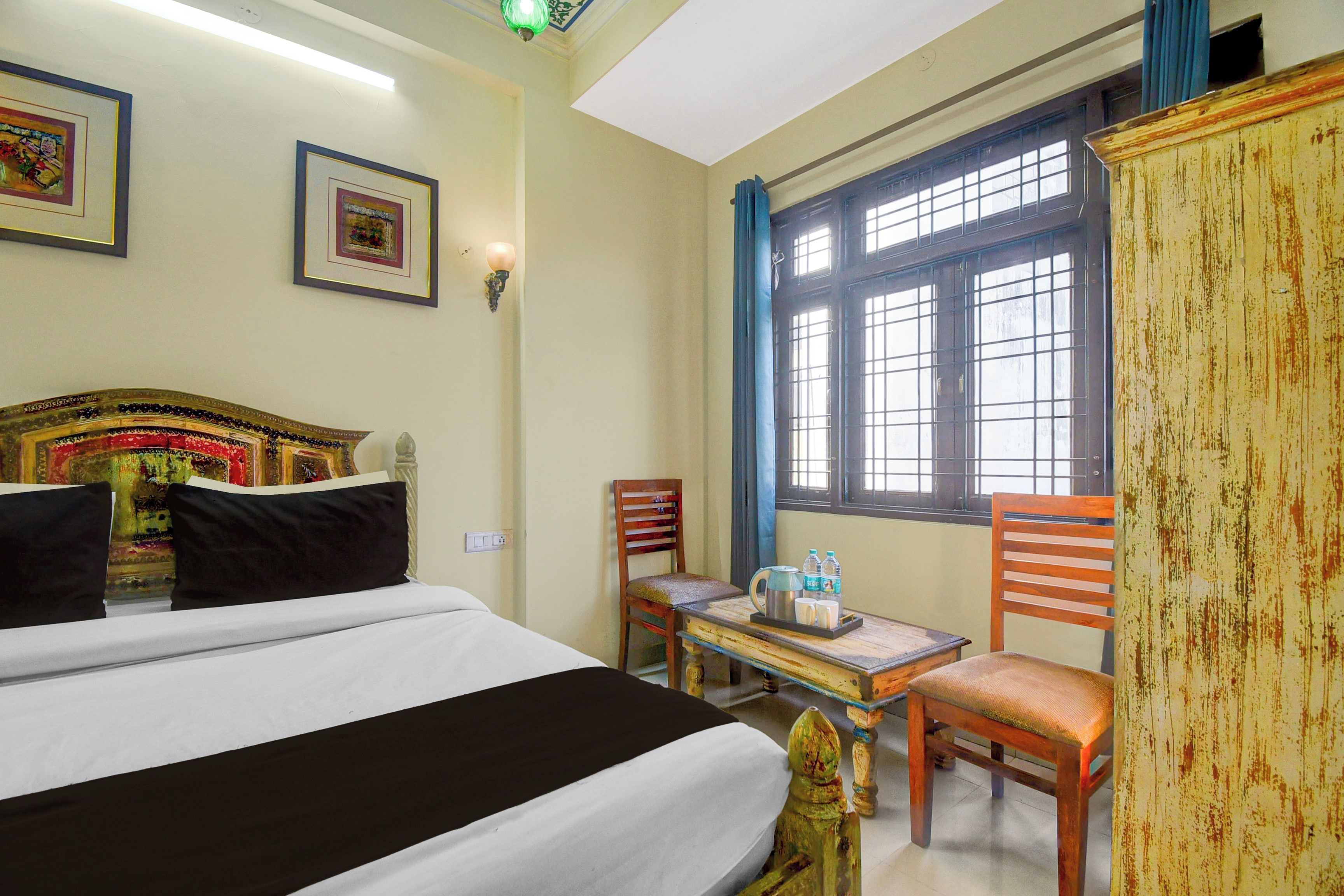 Una habitación de hotel con cama, dos sillas, una mesa con un servicio de botellas de agua, una lámpara y una ventana con cortinas.
