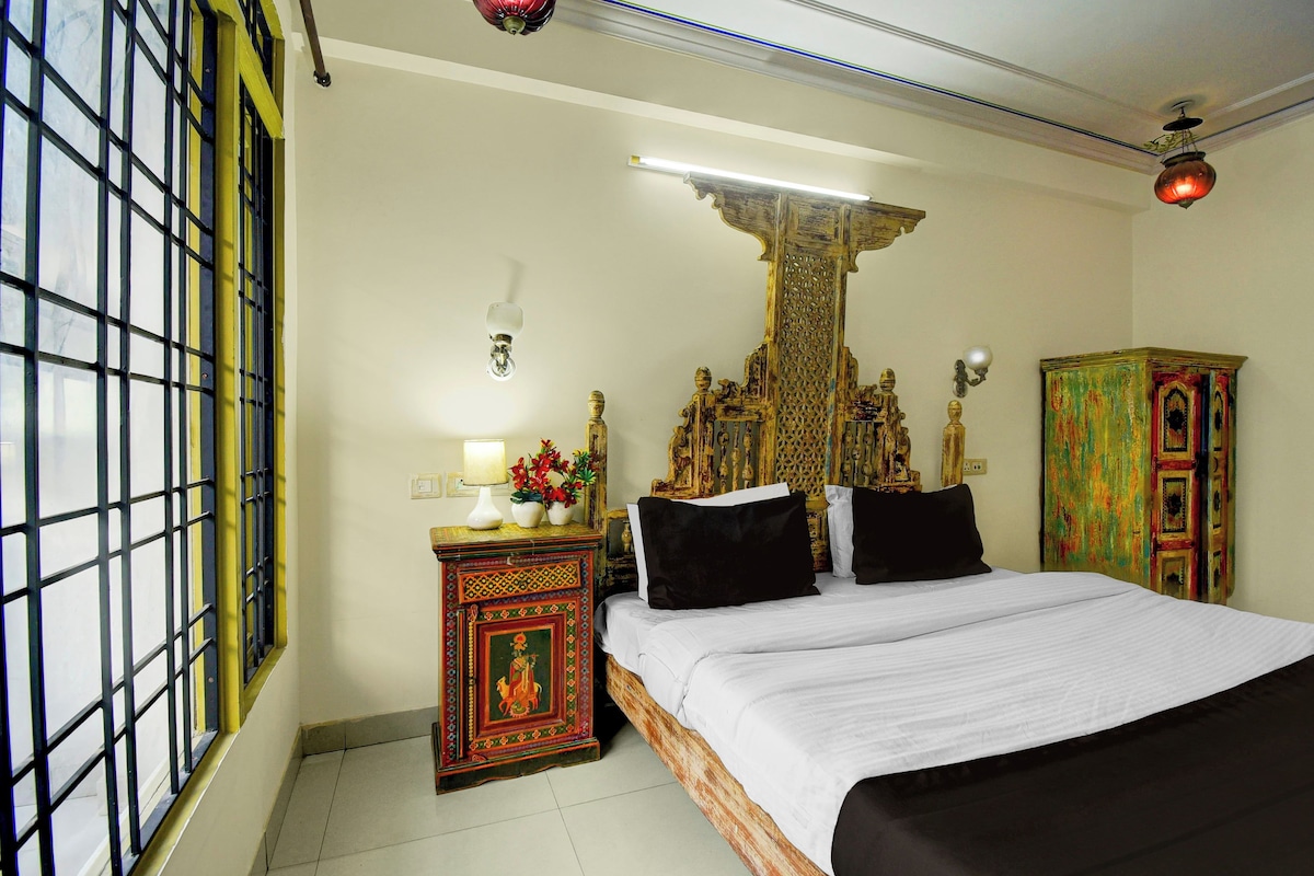 Habitació d'hotel amb un llit, una tauleta de nit amb una làmpada, una peça decorativa a la paret i una finestra amb reixes.