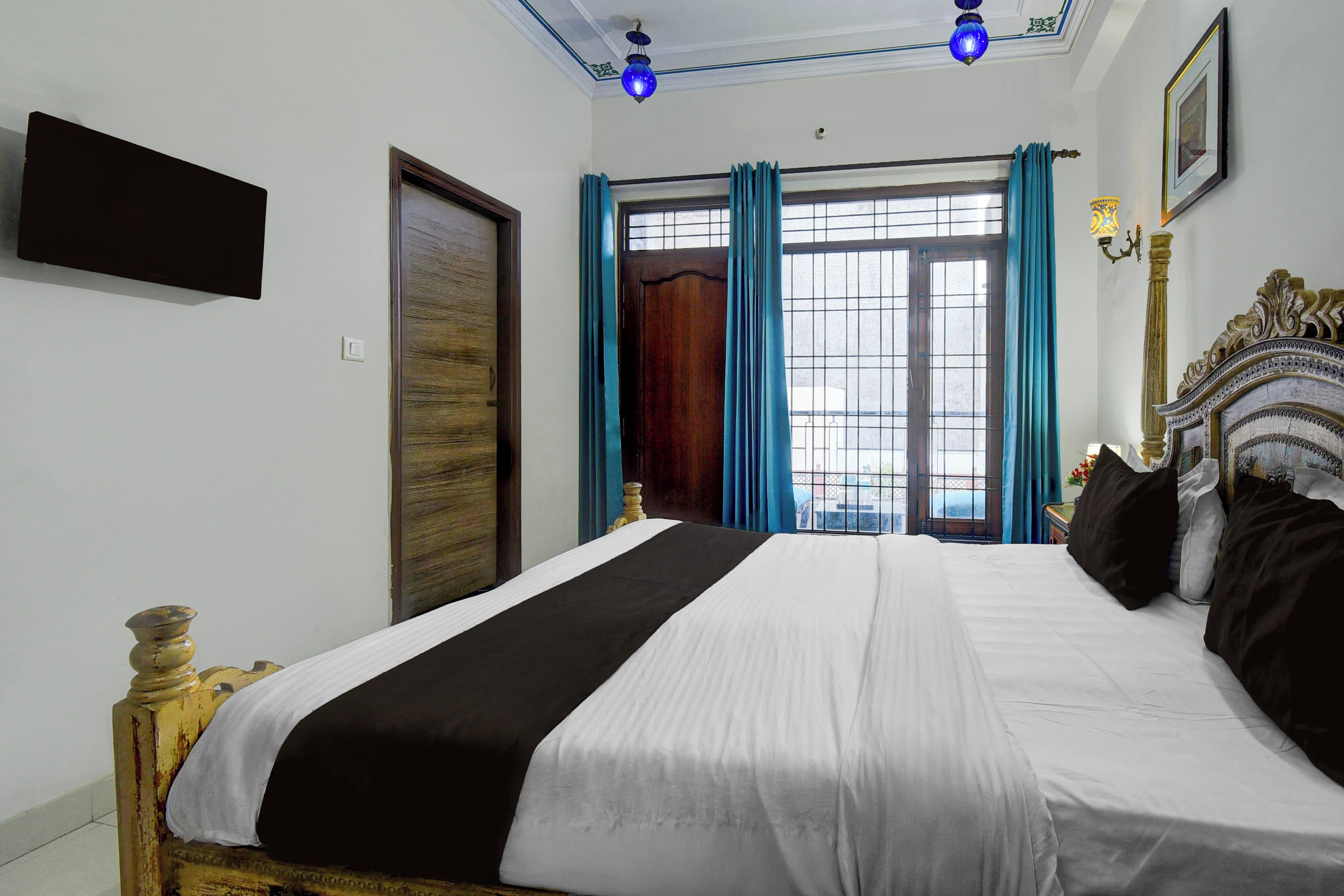 Una habitación de hotel con cama, puerta, ventana con cortinas y una lámpara fijada a la pared.
