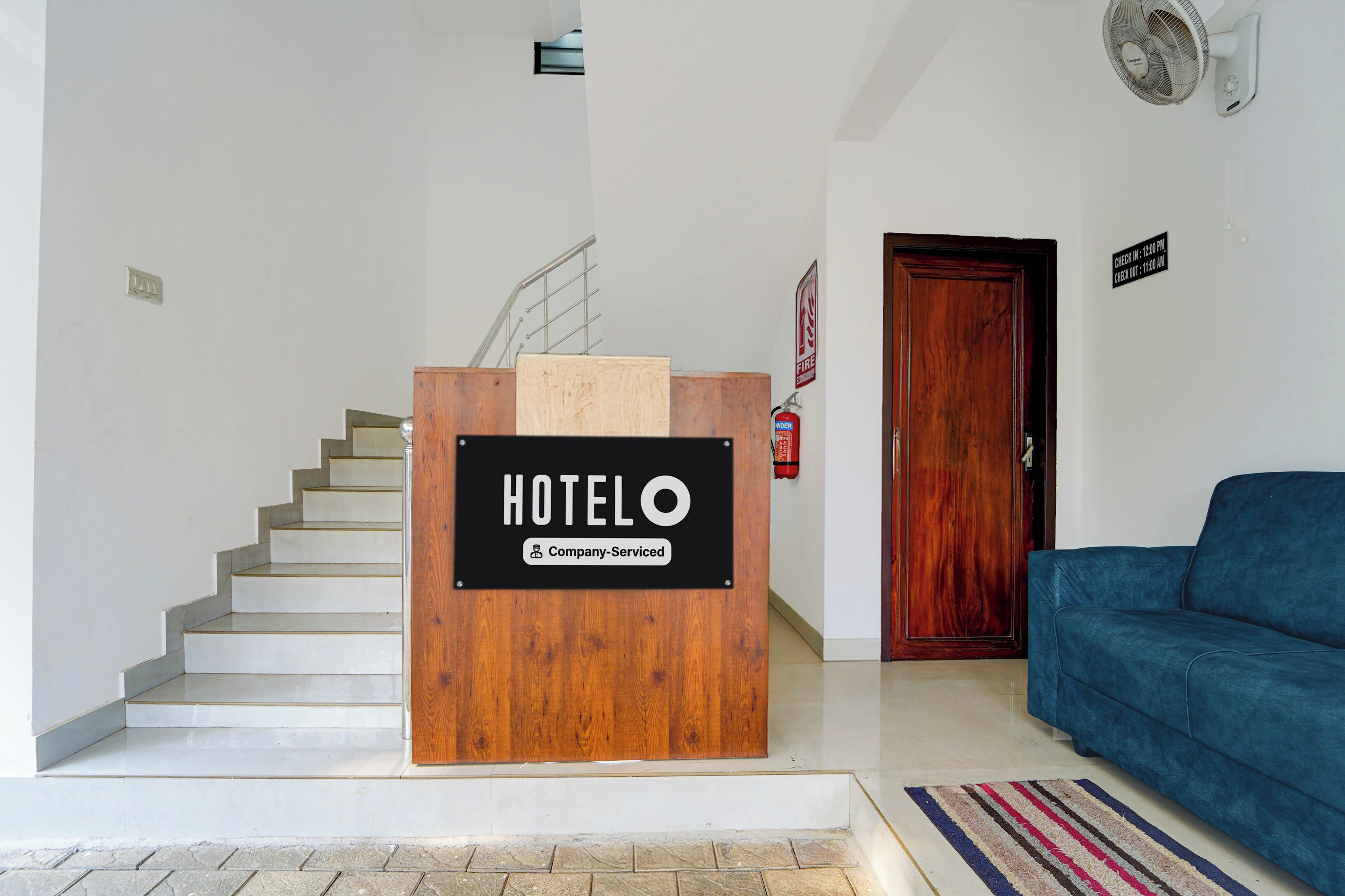 A hotel reception desk with a sign indicating it is company-serviced, a staircase, a wooden door, and a blue sofa in the living area.