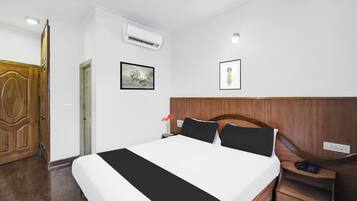 A hotel room with a bed, a nightstand, a wooden wardrobe, an air conditioning unit, a framed picture, and a wall-mounted light.