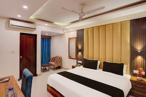 Habitación de hotel con cama, escritorio, silla, ventilador de techo y una ventana grande con cortinas.
