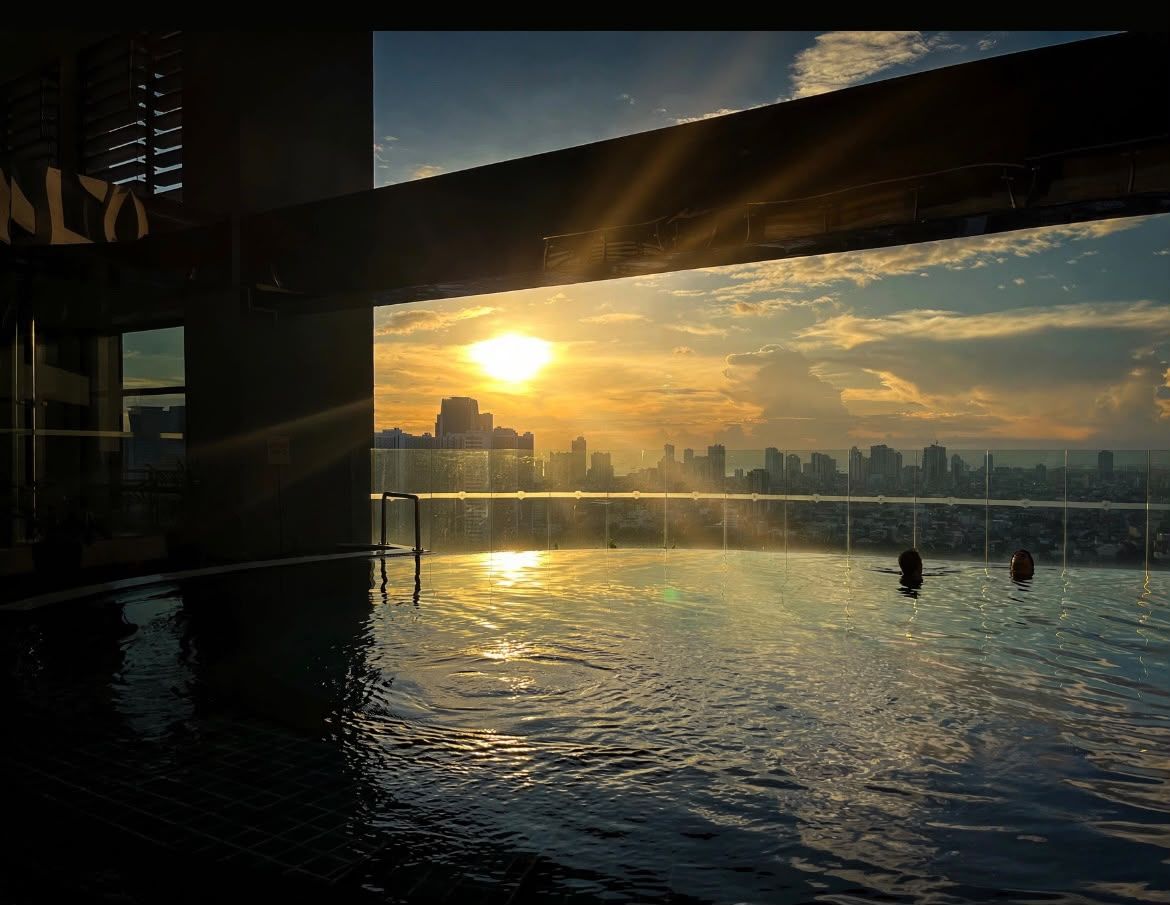 An indoor swimming pool with a city skyline view, a bridge structure, and a sun setting in the background.
