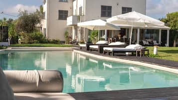 Une vue sur la piscine avec un fauteuil de relaxation, un bâtiment à la façade blanche et un espace extérieur aménagé avec un auvent.