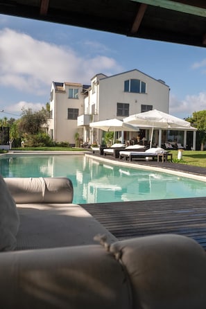 Blick auf ein Schwimmbecken mit Liegestuhl, ein Gebäude mit weißer Fassade und eine überdachte Sitzgelegenheit im Freien.