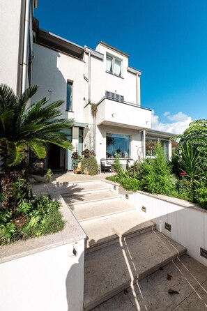 A modern two-story building with a flat roof, large windows, and a well-maintained garden with various plants and a palm tree. - Apartment 'Villa Verney' with Sea View, Wi-Fi and Air Conditioning (Albisola Superiore)