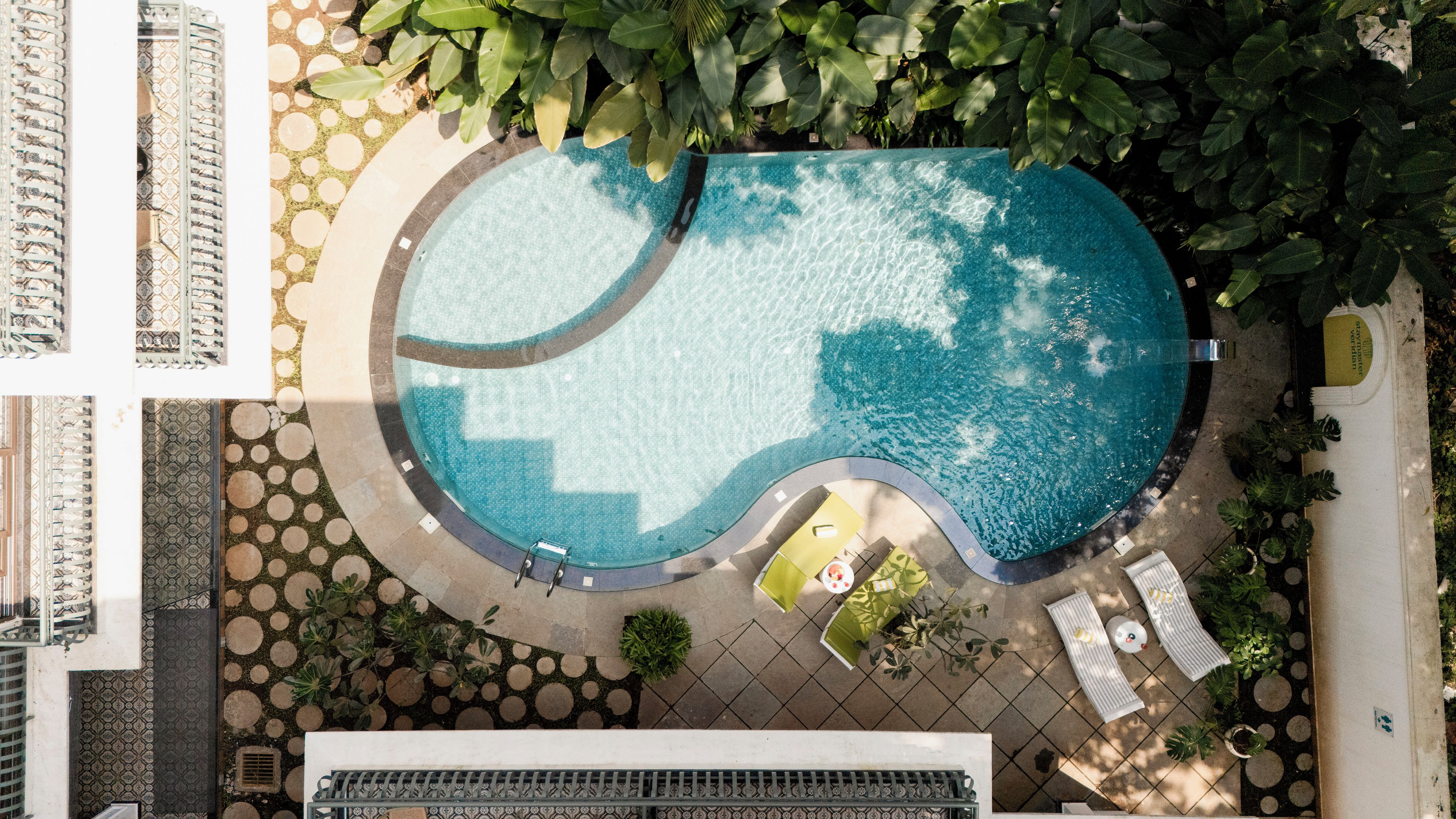 Vue aérienne d’une piscine à bordures courbes, entourée d’une terrasse carrelée et de verdure.