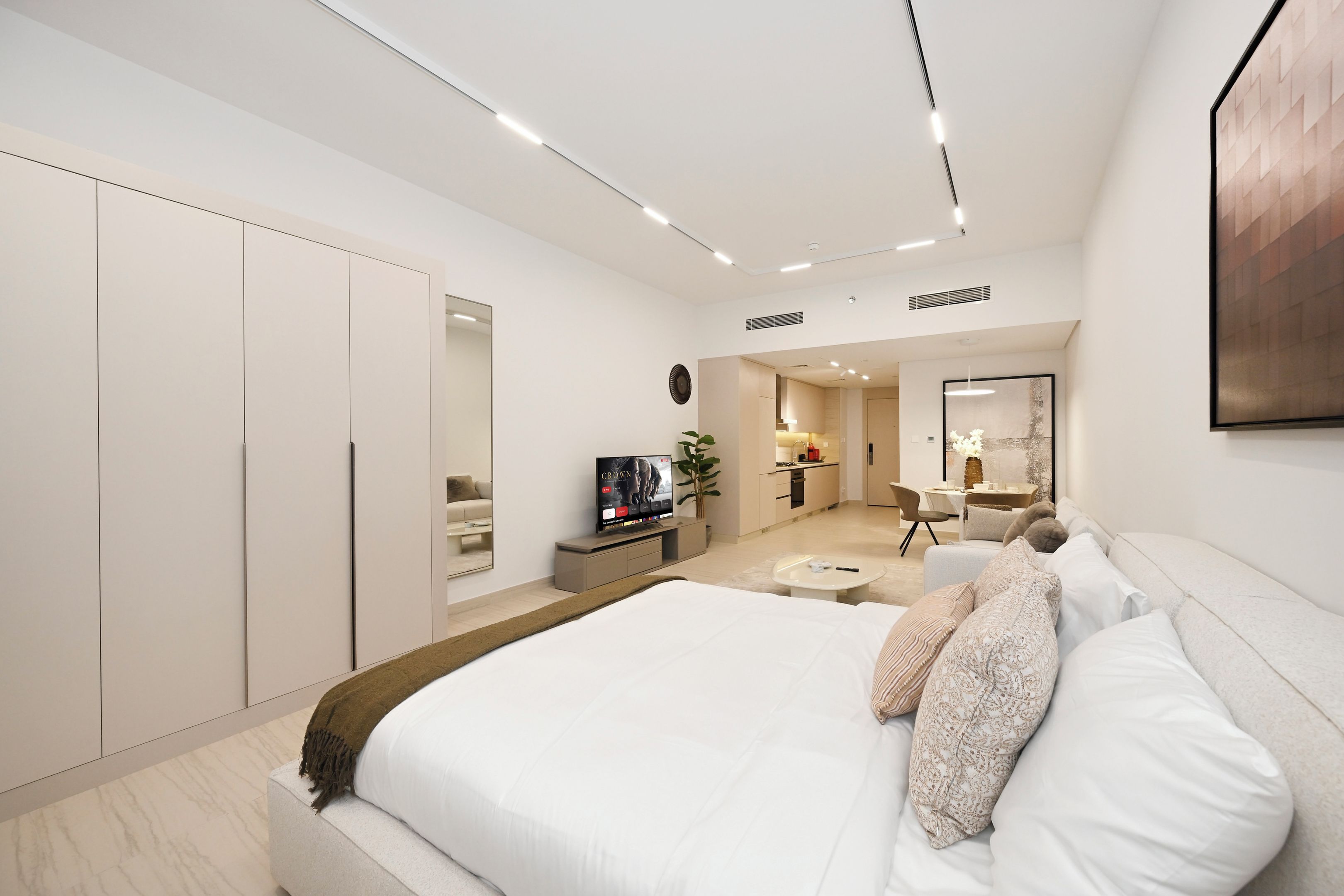 A modern bedroom with a large bed, a TV, a desk, and a kitchen area.