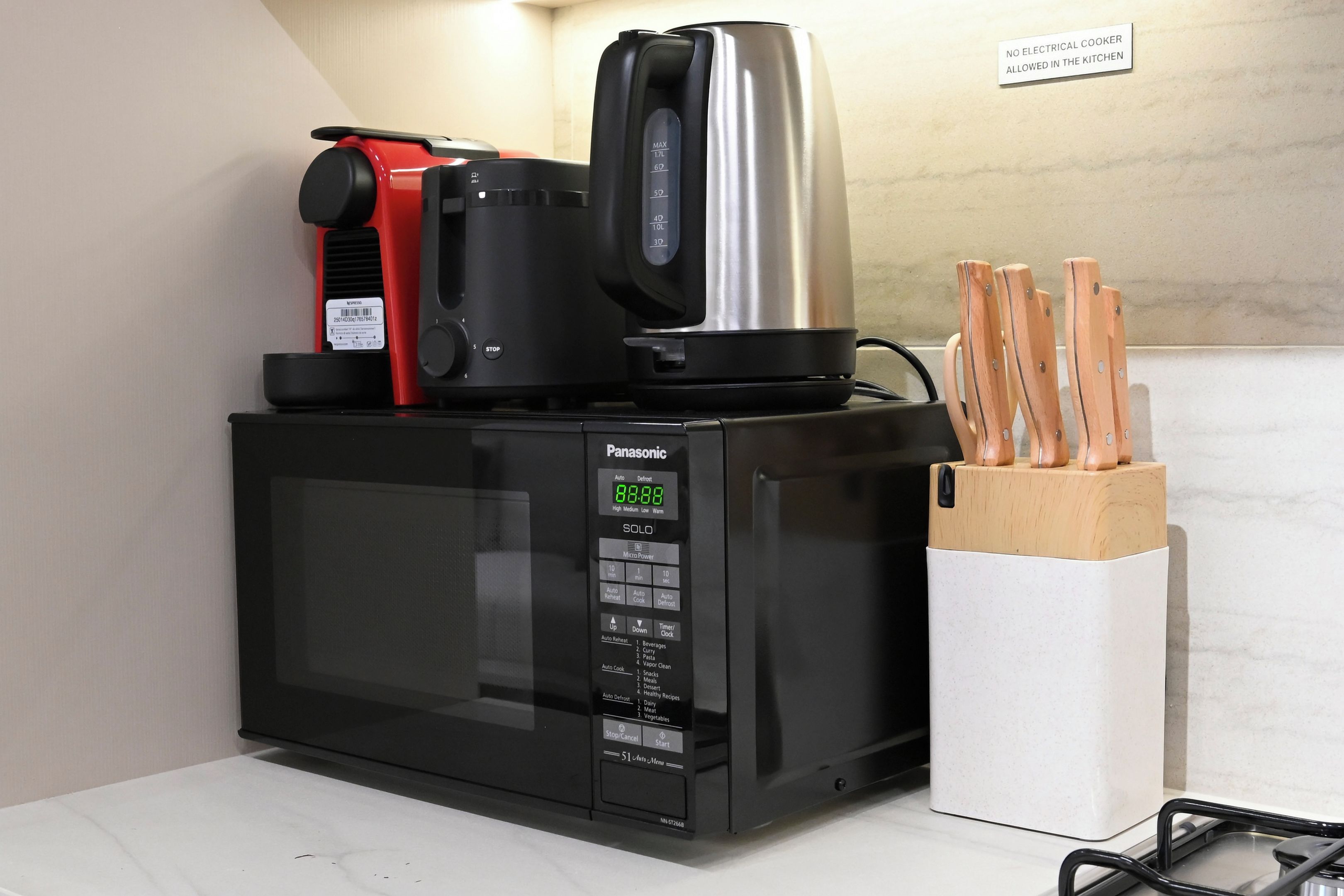 A black Panasonic microwave with a digital display, a red coffee maker, and a stainless steel kettle on top.