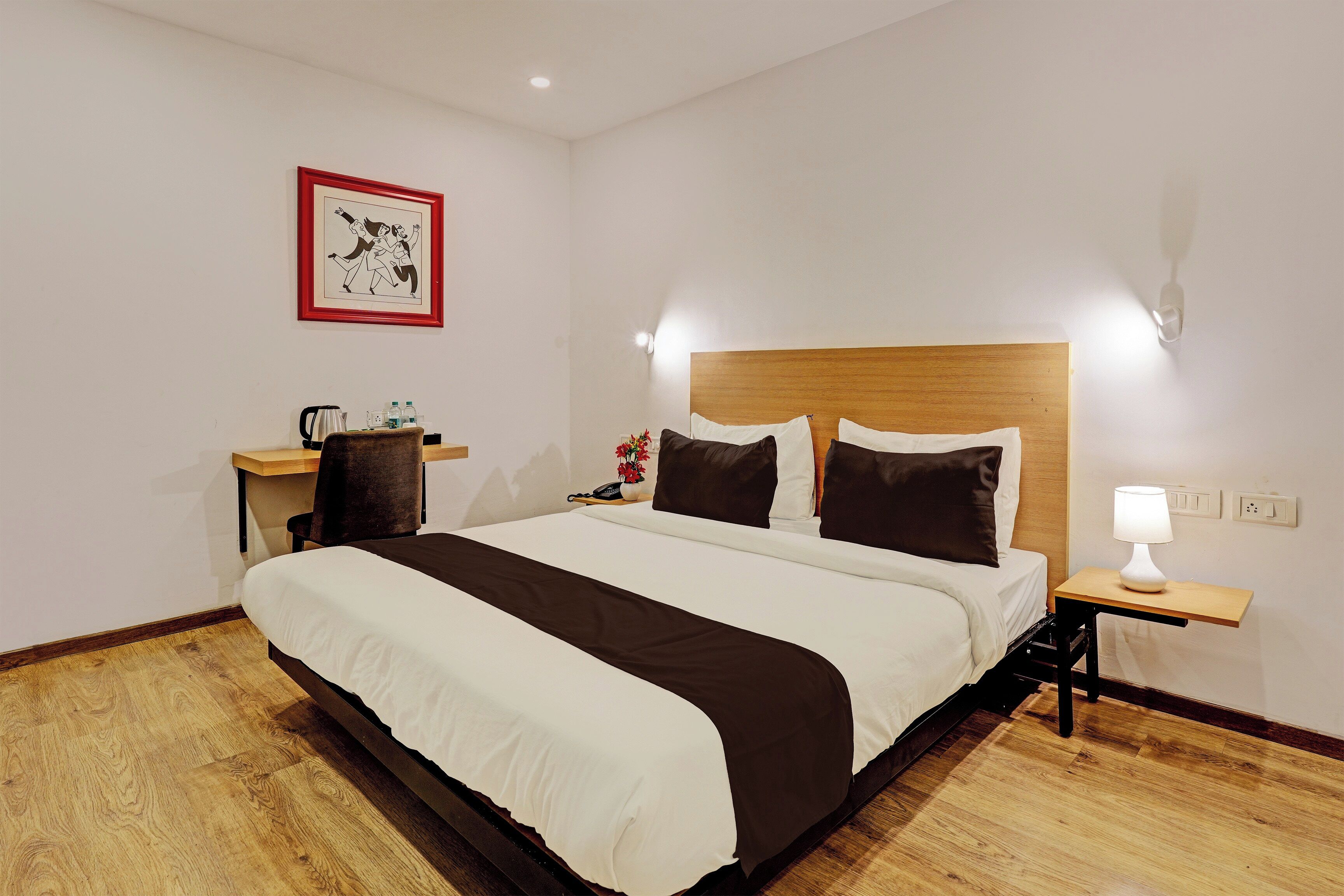 Una habitación de hotel con una cama grande, una mesita de noche con una lámpara, una silla, un escritorio y un cuadro en la pared.