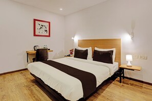 Una habitación de hotel con una cama grande, una mesita de noche con una lámpara, una silla, un escritorio y un cuadro en la pared.