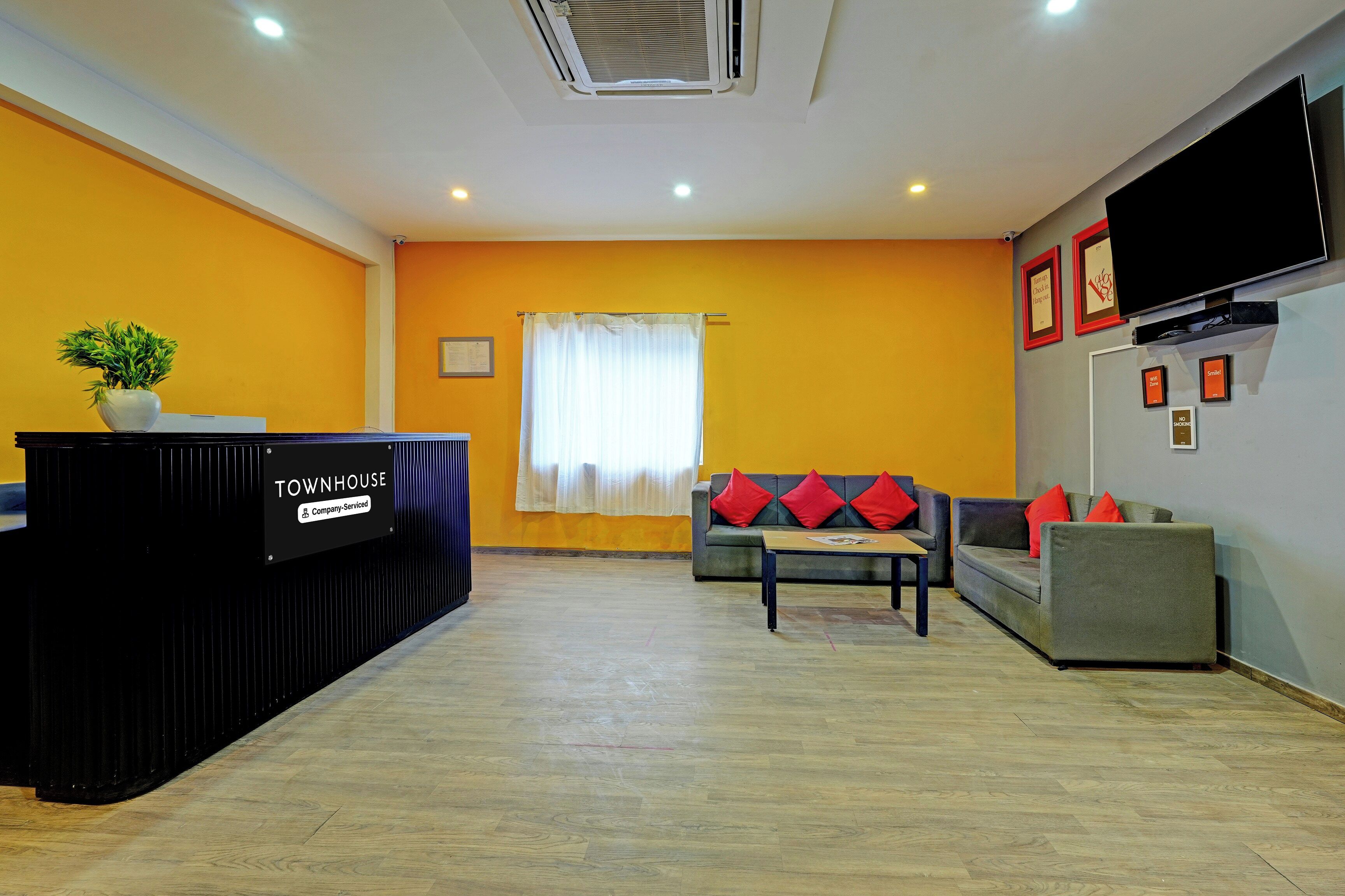 Zona de recepción con un mostrador negro identificado como 'TOWNHOUSE Company Serviced', un sofá con cojines rojos, un televisor montado en la pared y una ventana con cortinas blancas.