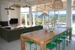 A dining area with a wooden table and green chairs, a sofa, and a television on a white cabinet. - Holiday Home 'Villa La Chanterelle' with Mountain View, Wi-Fi and Air Conditioning (Entrechaux)