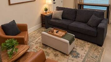 Una sala de estar con sofá, sillón, reposapiés y una mesita con una planta.