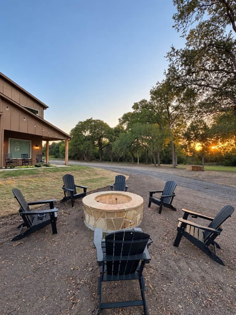 A circular outdoor seating area with chairs and a fire pit, surrounded by trees and a building with a covered patio.
