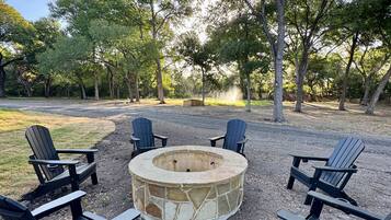 Un hogar circular de piedra rodeado por cuatro sillas Adirondack en un entorno exterior con árboles y cielo despejado.