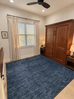 Een kamer met een groot blauw tapijt, een houten garderobe, een raam met gordijnen en een plafondventilator.