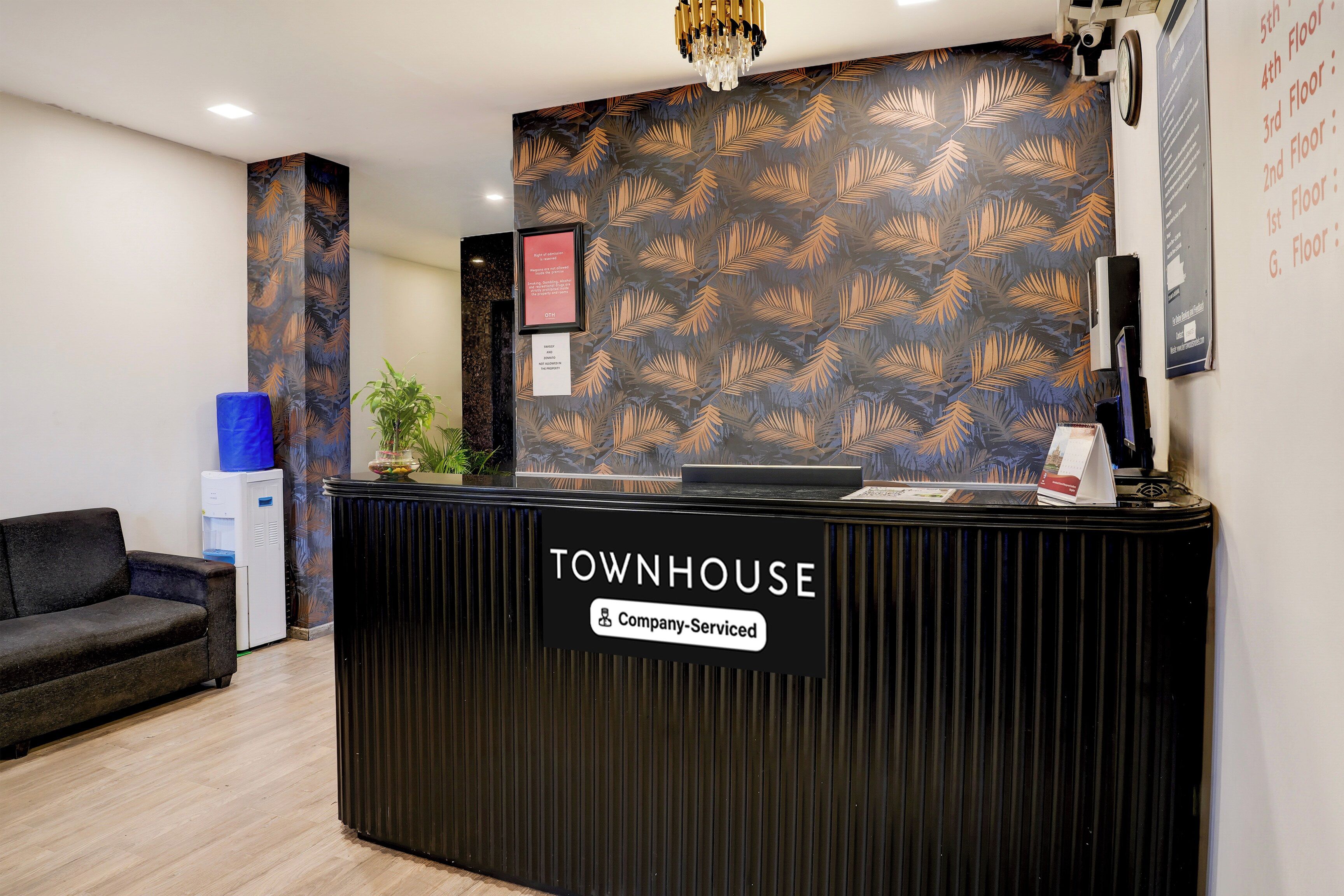 Mostrador de recepción con la etiqueta 'TOWNHOUSE', un cartel que dice 'Servicio de Empresa', una planta en maceta, un enfriador de agua azul y un sofá negro.