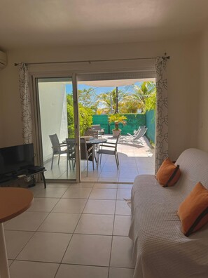 A room with a bed, a TV, a dining table with chairs, and a sliding glass door leading to an outdoor area with a patio and greenery. - Caribbean Getaway in Le Gosier, Sea View • Pool • WiFi • Near Restaurants (Le Gosier)