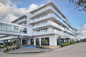 A modern multi-story hotel with a glass facade, outdoor seating area, and a clear sky. - RentPlanet - Apartamenty Wydmowa (Kołobrzeg)