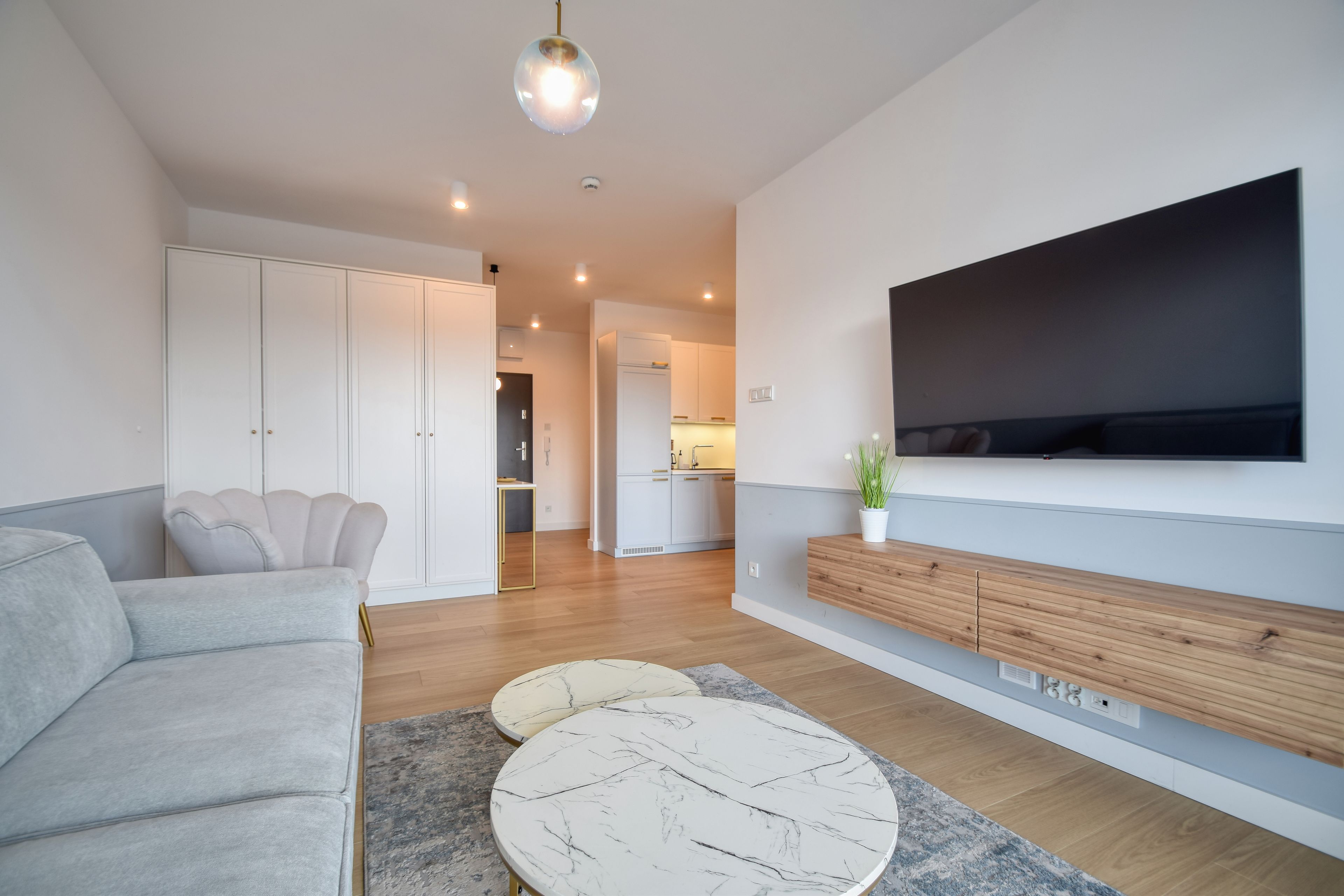 A modern living room with a grey sofa, a marble coffee table, a flat-screen TV mounted on the wall, and a wooden cabinet below.