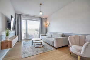 Ein modernes Wohnzimmer mit einer Couch, einem Sessel, einem Fernseher und Blick durchs Fenster.