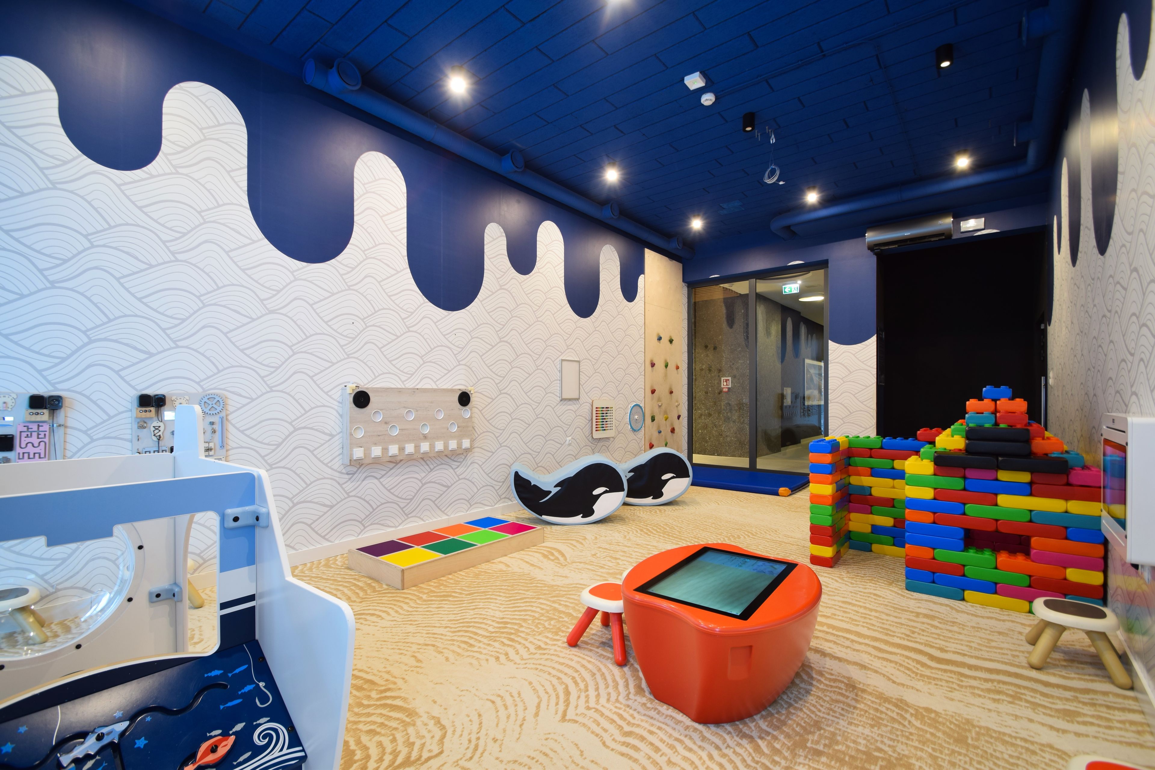 A children's play area with a climbing wall, colorful building blocks, and a tablet on a stool.