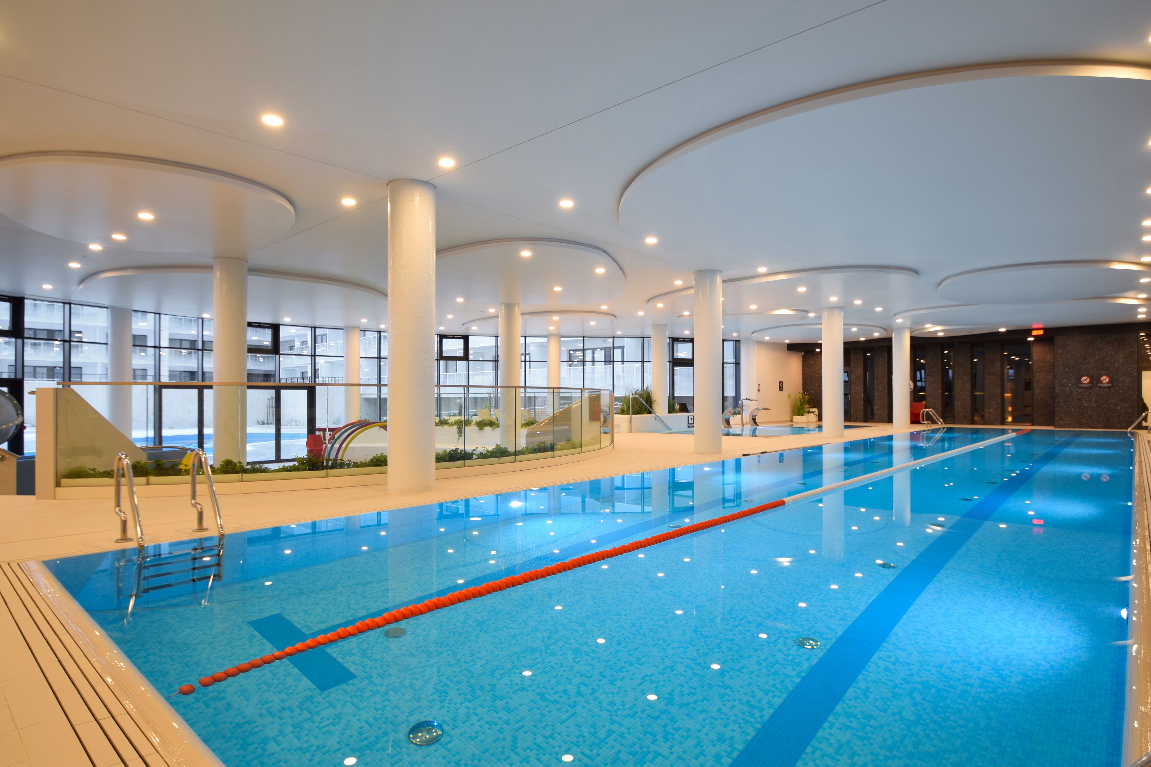 An indoor swimming pool with clear lanes, surrounded by large windows and columns.