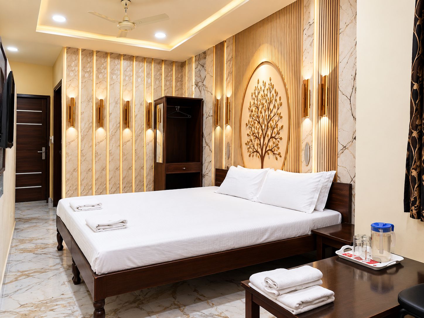 Une chambre d’hôtel avec un grand lit, une armoire, un miroir, une décoration murale en forme d’arbre et une table de chevet sur laquelle se trouve un plateau avec des serviettes.