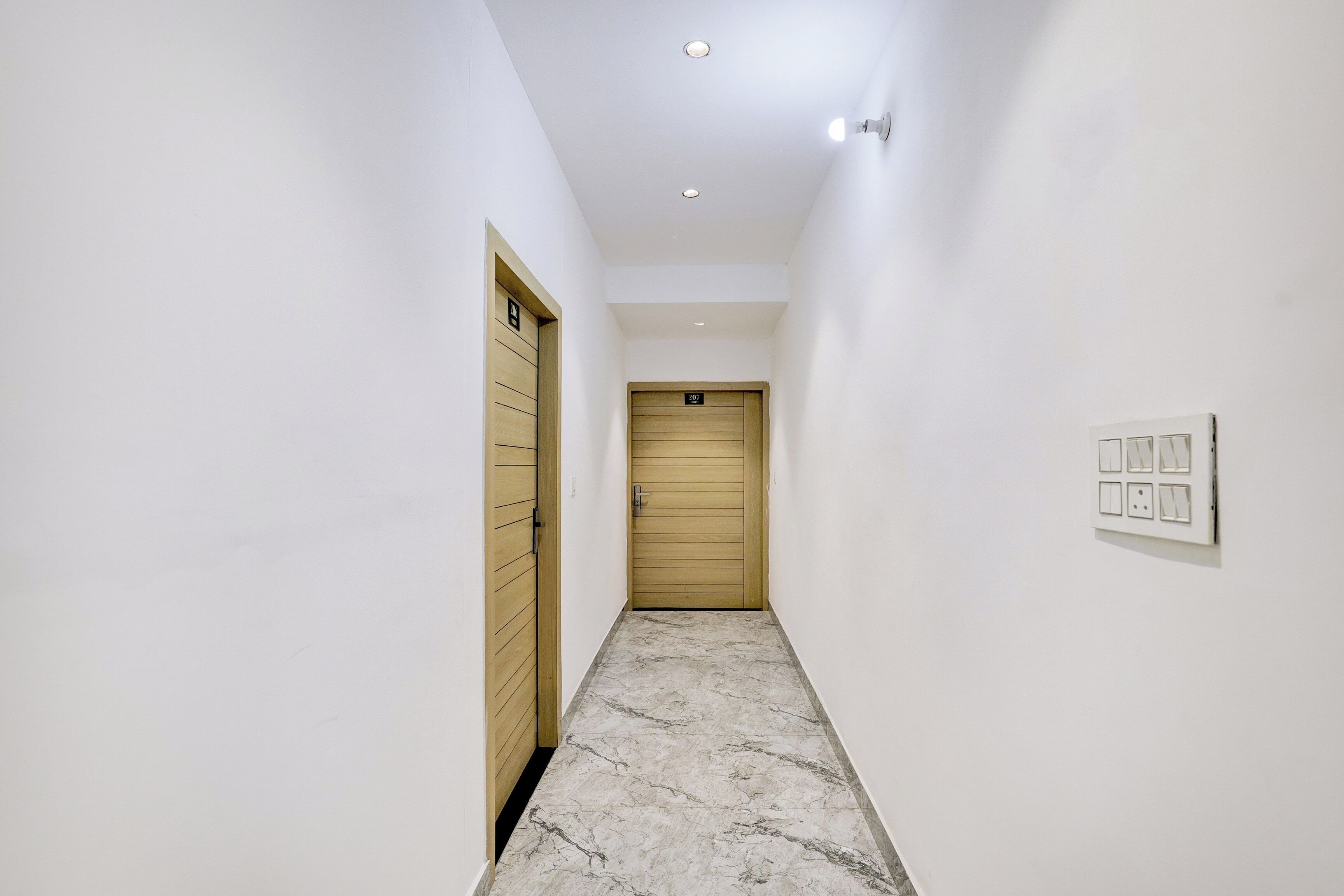 Un pasillo con una puerta de madera al final, un interruptor de luz en la pared y un piso de mármol.