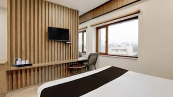 A hotel room with a bed, a TV mounted on a wooden panel wall, a small table with a kettle and cups, a chair, and a window with a view of buildings.