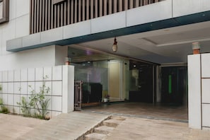 Entrada de un edificio moderno con puerta de vidrio, un escalón y una planta en maceta a la izquierda.
