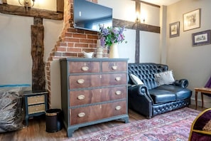 Een kamer met een open haard van baksteen, een donker houten dressoir met lades, een zwarte leren bank en een kamerkleed met een patroon.